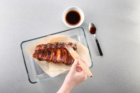 Woman adding soy sauce to roasted pork ribs in baking tray on tableの写真素材