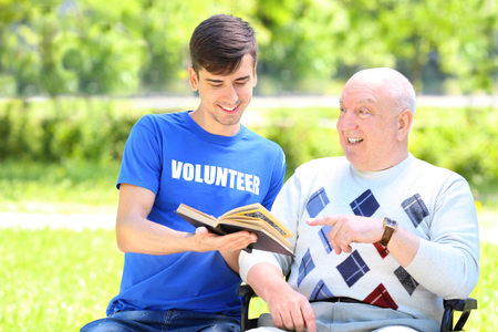 Young male volunteer reading book to senior man on wheelchair outdoorsの写真素材