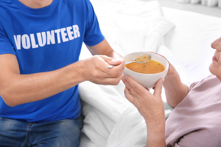 Young male volunteer feeding elderly woman in light roomの写真素材
