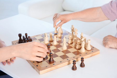 Young female volunteer playing chess with elderly woman in light room