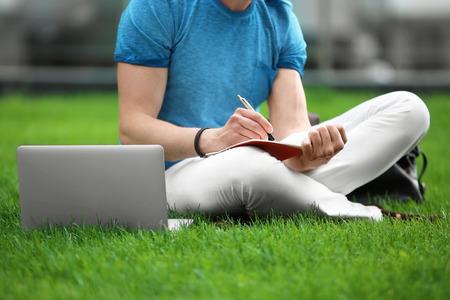 Young blogger with laptop sitting on green grass, outdoors