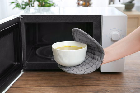 Woman taking bowl with delicious soup out of microwave in kitchenの写真素材