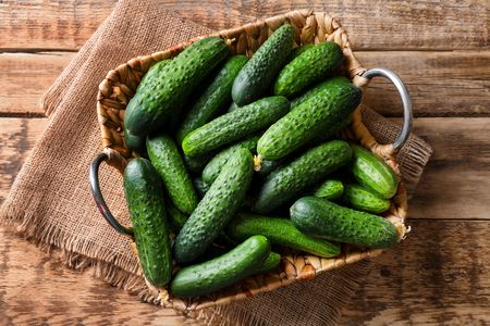 Many green fresh cucumbers in wicker basket on wooden tableの写真素材