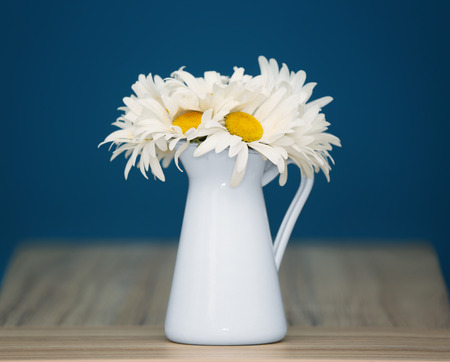Beautiful chamomiles in white jug on wooden table against color backgroundの写真素材