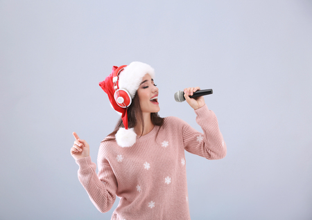 Beautiful young woman in Santa hat singing Christmas songs on light backgroundの写真素材