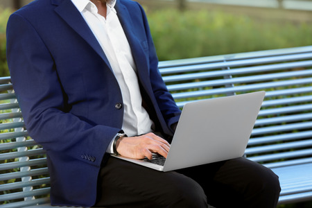 Stylish young man with laptop on bench, outdoorsの写真素材