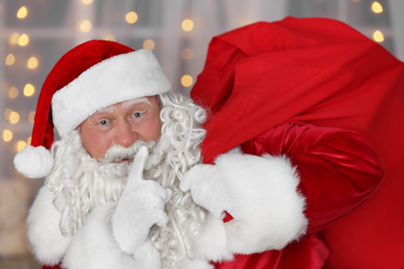 Authentic Santa Claus with big red bag full of gifts against blurred Christmas lightsの写真素材