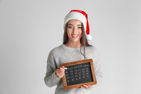Young woman in Santa hat with chalkboard counting days until Christmas, on light backgroundの写真素材
