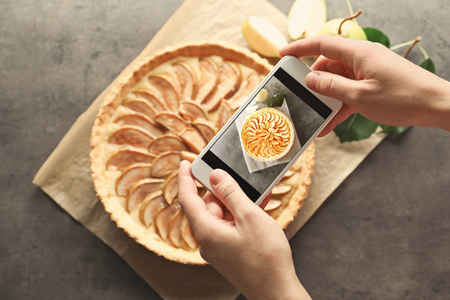 Woman taking photo of tasty pear tart with mobile phone in kitchenの写真素材