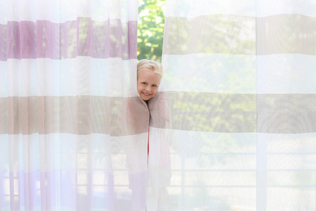 Cute little girl hiding behind curtains at homeの写真素材