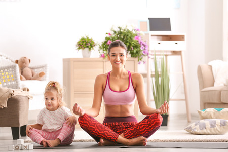 Beautiful young woman with little daughter practicing yoga at homeの写真素材