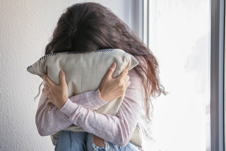 Sad young woman with pillow sitting near windowの写真素材