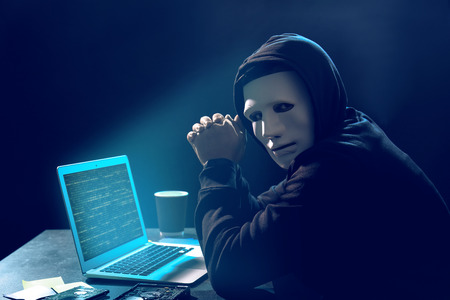 Hacker in mask with laptop in dark room