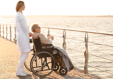 Young nurse from care home and senior woman in wheelchair near riverの写真素材