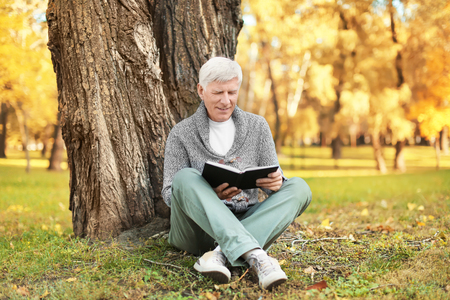 Stylish mature man reading book while sitting near tree in autumn parkの写真素材