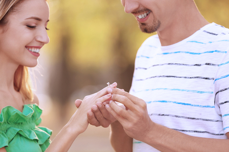 Young man putting engagement ring on fiancee's finger in parkの写真素材
