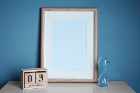 Empty frame, wooden calendar and hourglass on table near white wall