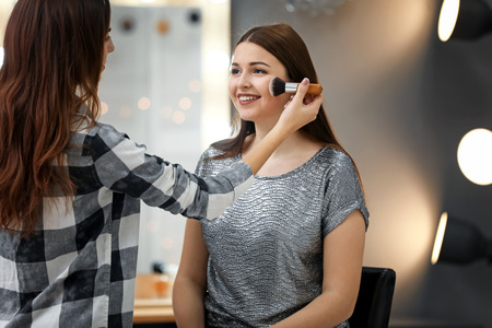Visagiste applying makeup onto face of beautiful young woman in roomの写真素材
