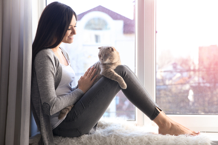 Young woman with cute pet cat sitting on windowsill at homeの写真素材