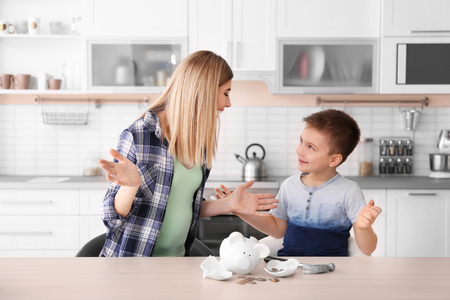 Mother and little son with broken piggy bank on table at homeの写真素材