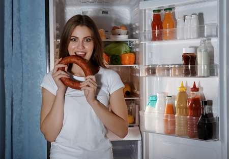 Young woman with sausage standing near refrigerator at nightの写真素材