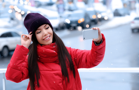 Beautiful young woman taking selfie with mobile phone outdoors on winter day