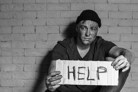 Homeless poor man holding carton board with word HELP while sitting near brick wallの写真素材
