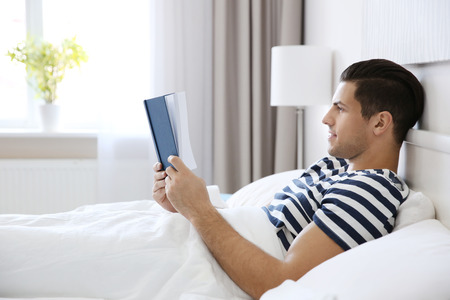 Young handsome man reading book in bed at home