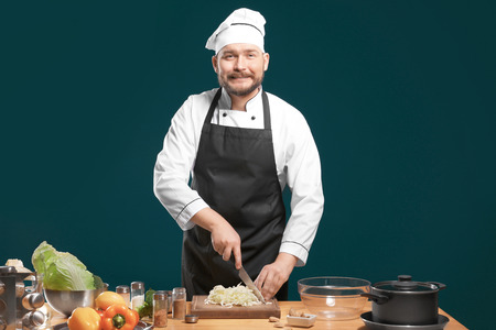 Handsome male chef cutting cabbage at table against color background