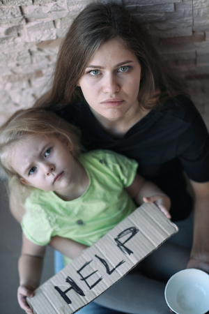 Homeless poor woman and her little daughter sitting near brick wall and asking for helpの写真素材