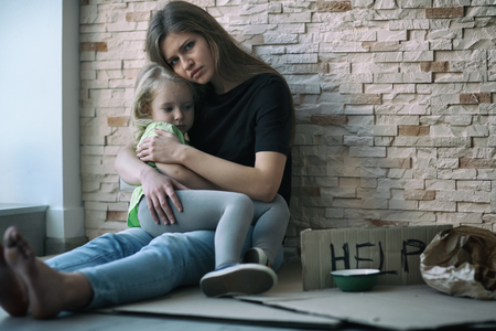 Homeless poor woman and her little daughter sitting near brick wall and asking for help