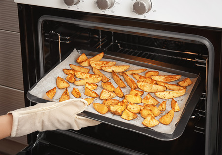 Woman taking baking sheet with delicious potatoes from ovenの写真素材