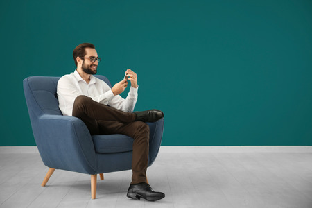Handsome man with mobile phone sitting in comfortable armchair against color wallの写真素材