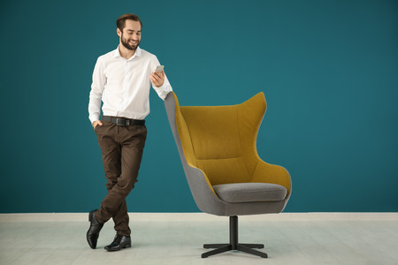 Handsome man with mobile phone standing near comfortable armchair against color wallの写真素材