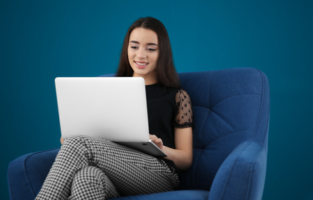 Beautiful young woman with laptop sitting in comfortable armchair on color backgroundの写真素材