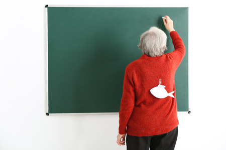 Senior teacher with paper fish on her back writing on chalkboard. April fool's day celebrationの写真素材