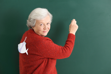 Senior teacher with paper fish on her back writing on chalkboard. April fool's day celebrationの写真素材