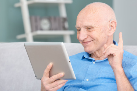 Senior man with hearing aid using tablet computer indoorsの写真素材