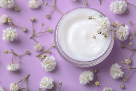 Jar of body cream and beautiful flowers on color background