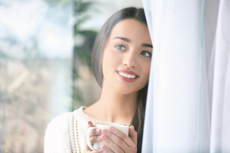 Beautiful young woman with cup of hot tea at home, view through windowの写真素材