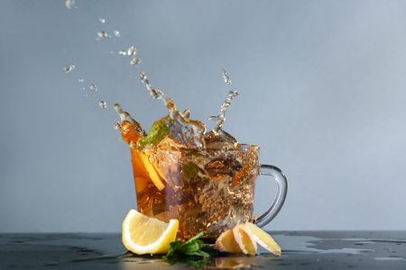 Glass cup and splash of delicious lemon tea with ginger on table against color backgroundの写真素材