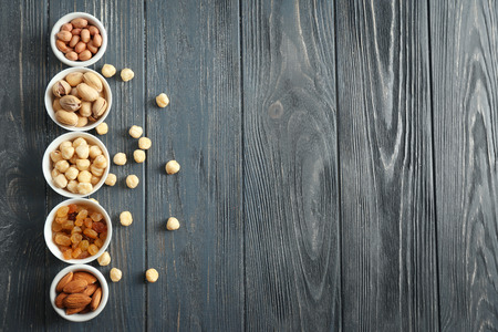 Different kinds of nuts and raisins in bowls on wooden background, top view