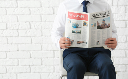 Businessman sitting on chair while reading newspaper against brick wall backgroundの写真素材