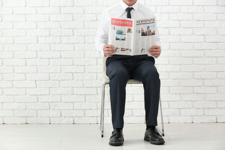 Businessman sitting on chair while reading newspaper against brick wall indoorsの写真素材