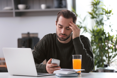 Male freelancer using smartphone while working with laptop in home officeの写真素材