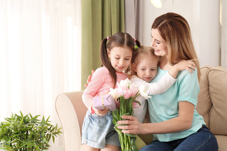 Mother receiving gift and flowers from her cute little daughters at homeの写真素材