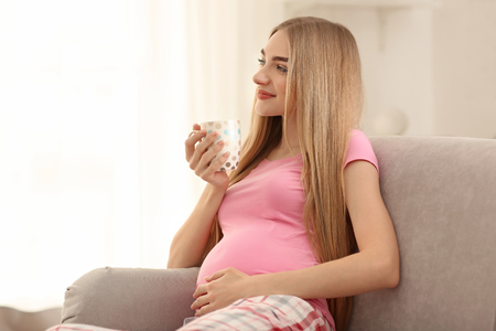 Young pregnant woman drinking tea on sofa at homeの写真素材