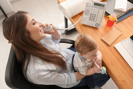 Young mother with baby talking on telephone while working in officeの写真素材