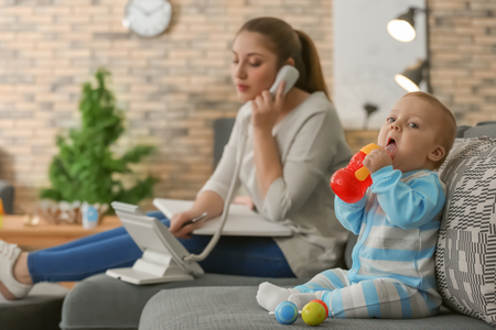 Cute little baby sitting on sofa while mother working at homeの写真素材