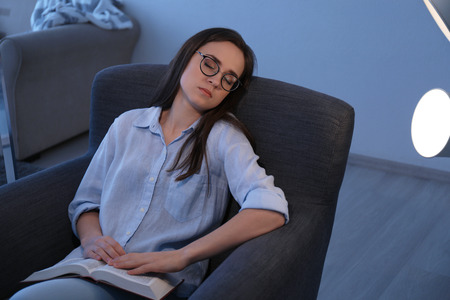 Tired young woman with book sleeping on armchair at home in evening